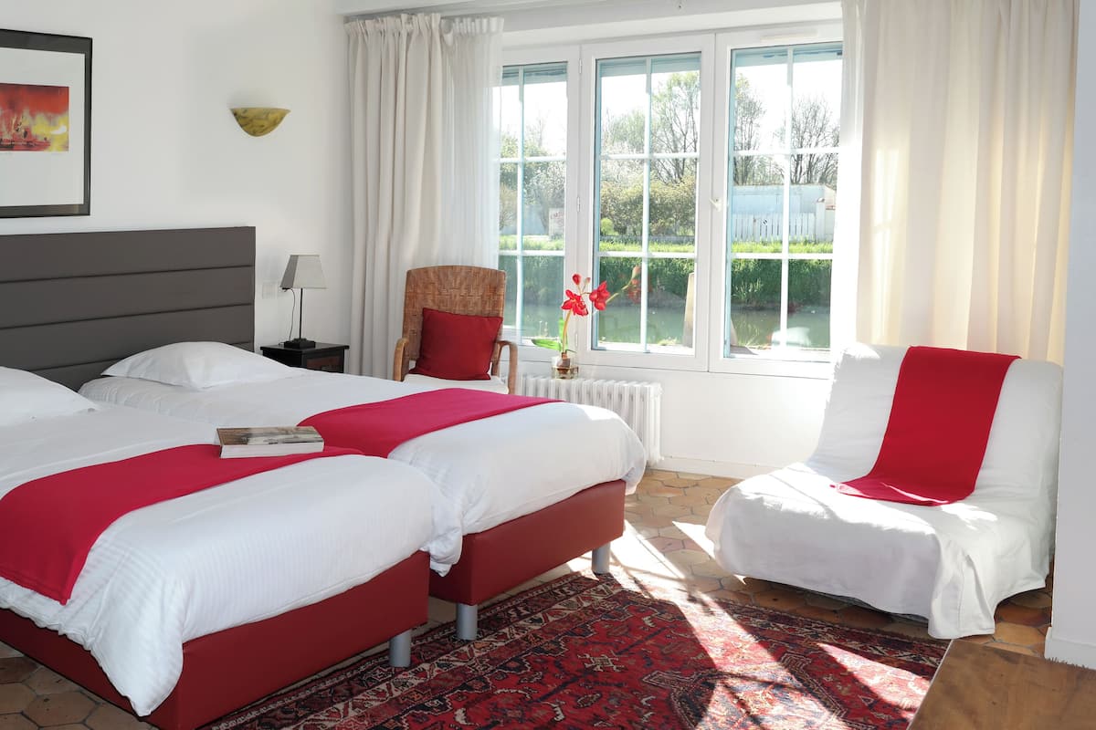 Habitación de hotel con dos camas, una silla roja, un ventanal con vistas a zonas verdes y un cuadro en la pared.