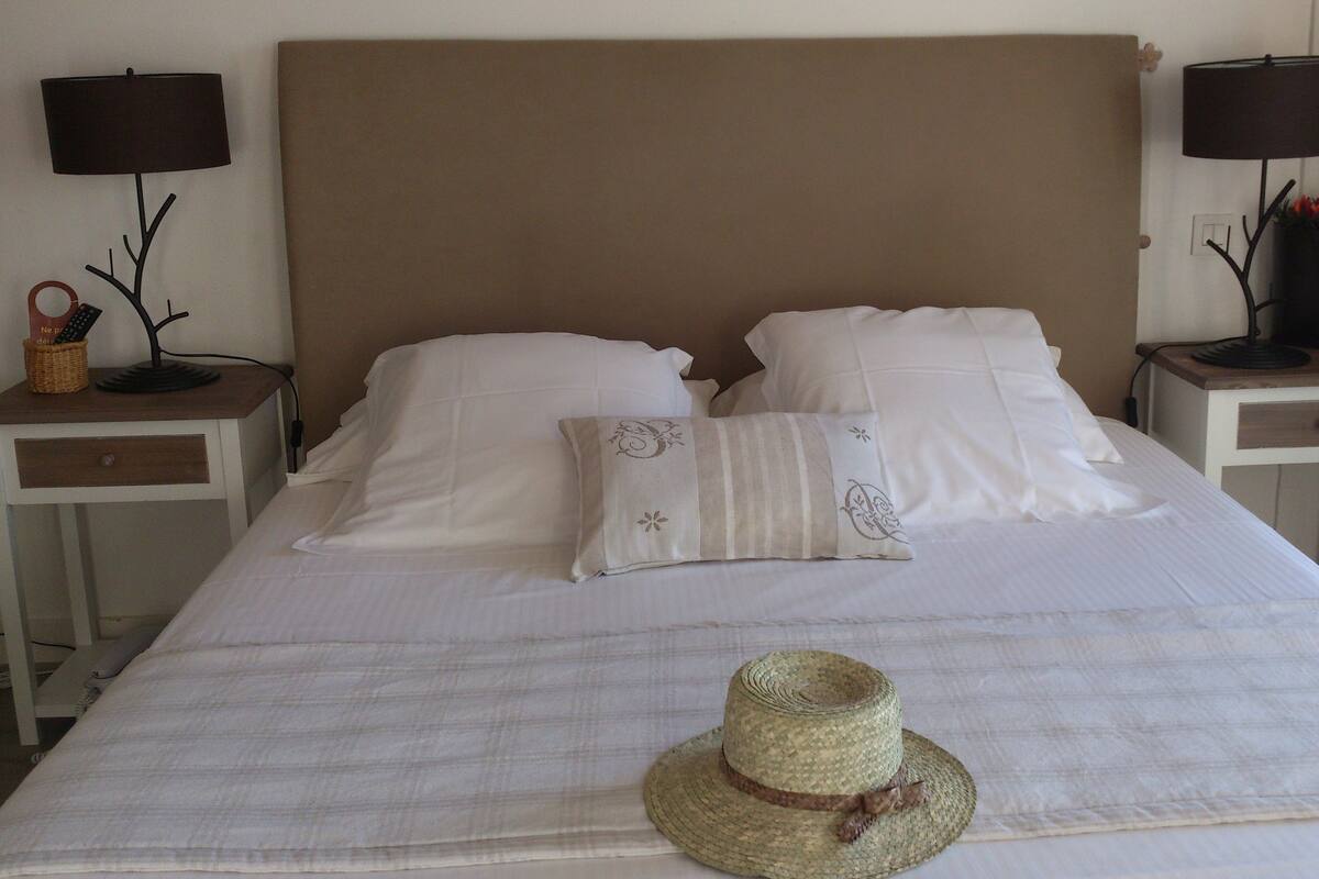 Una cama bien hecha con sábanas blancas, dos mesitas de noche con lámparas y un sombrero tejido sobre la cama.