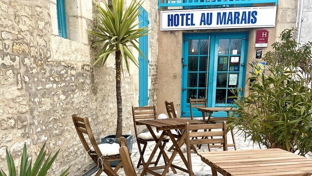 Mobiliario de madera exterior con cojines, suelo de piedra y un edificio con un letrero que dice 'HOTEL AU MARAIS'.