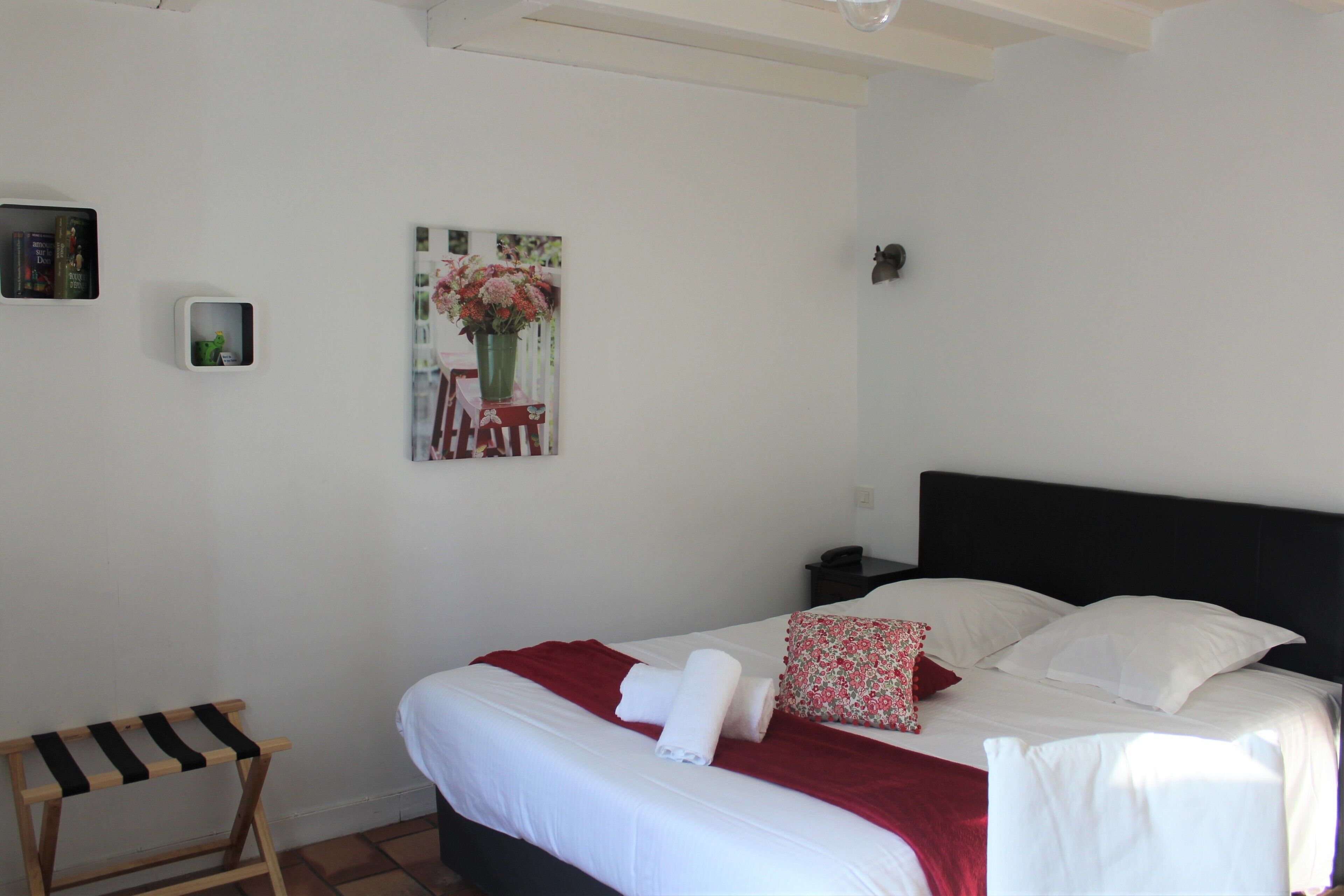 A neatly made bed with white linens and a red blanket, a small bedside table with a lamp, and a painting of flowers on the wall.