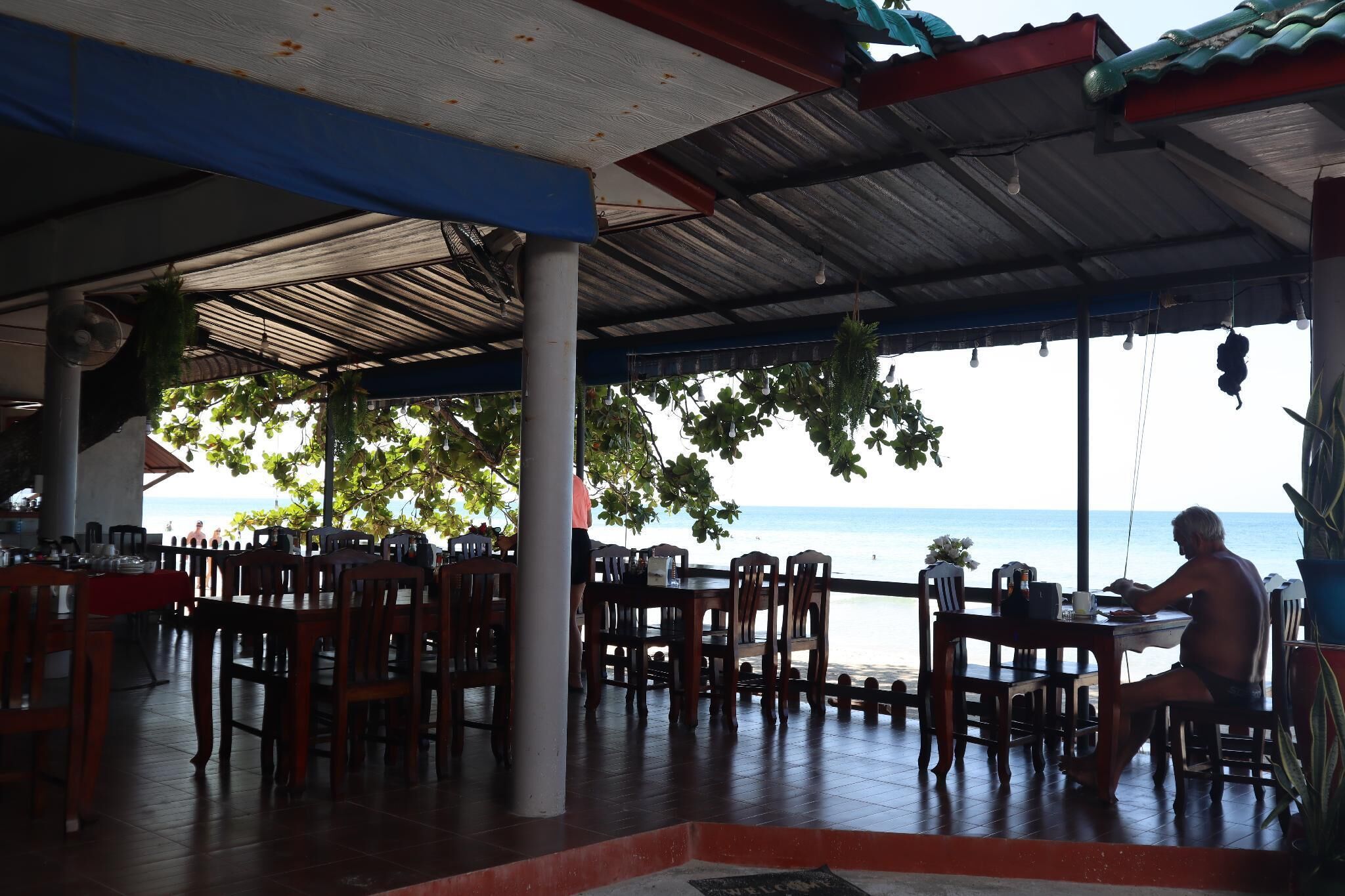 A covered outdoor dining area with wooden tables and chairs, overlooking a beach.