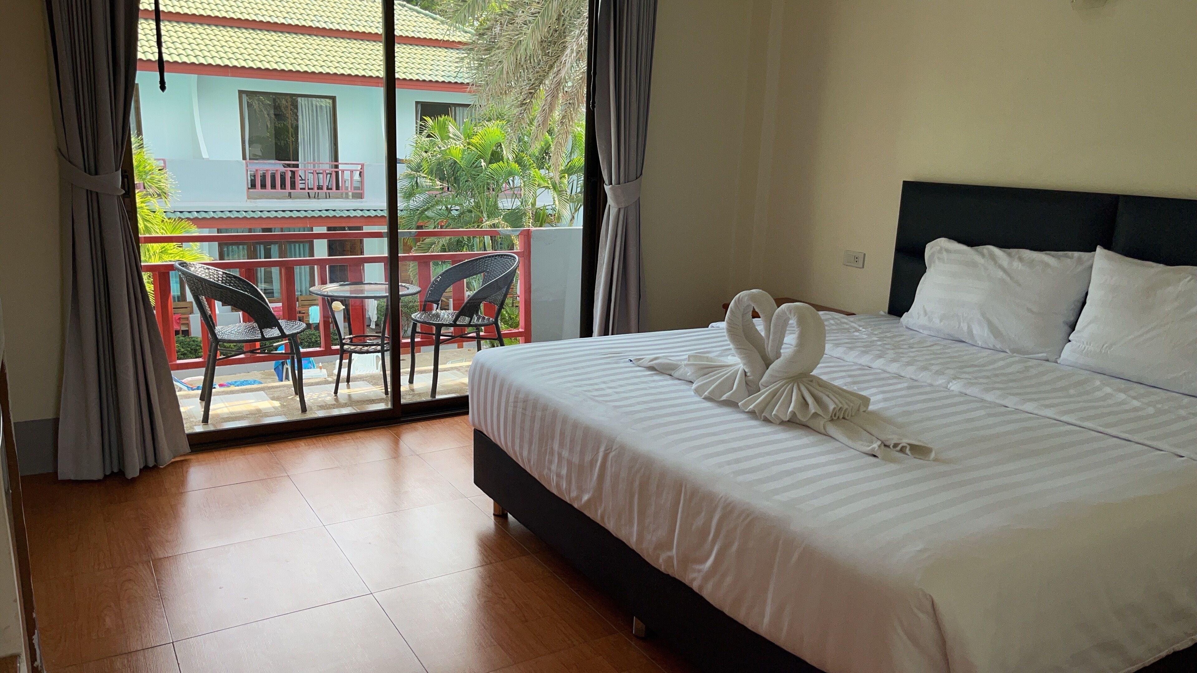 A hotel room with a bed, a balcony with chairs, and a view of a building with a red roof.