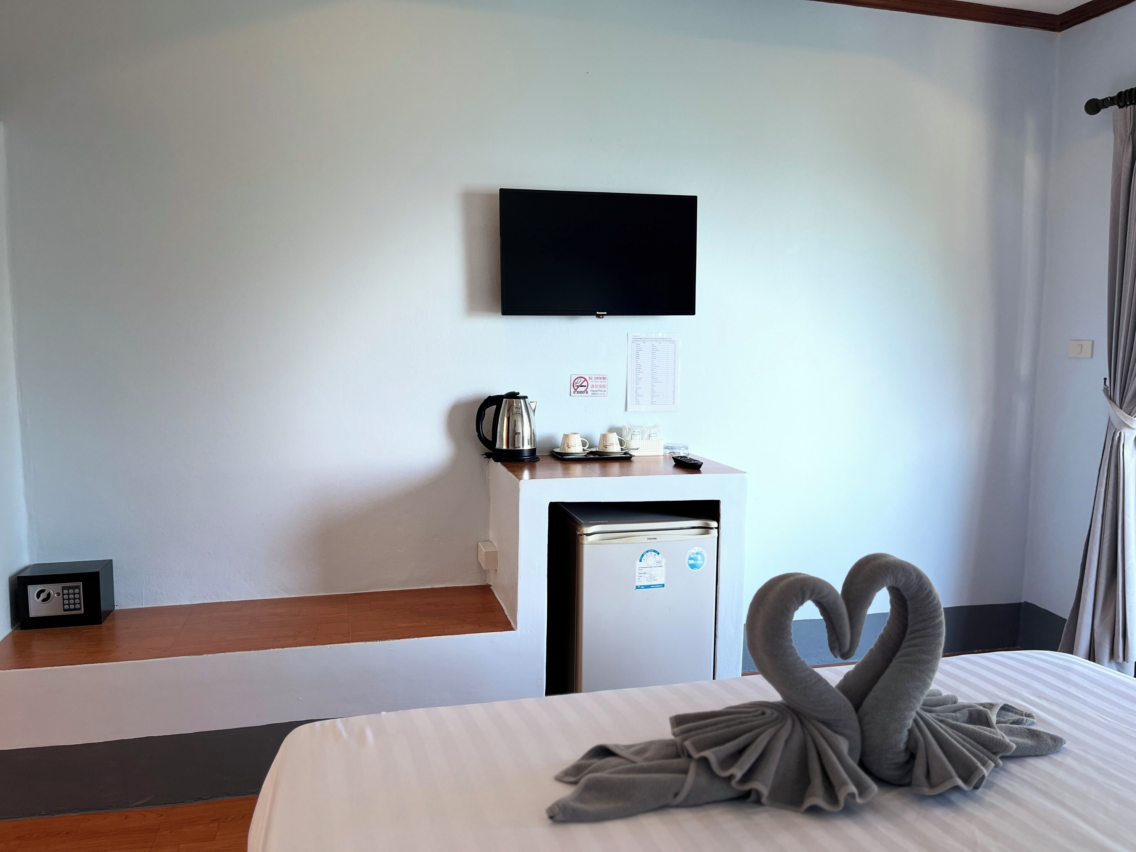 A hotel room with a bed, a small desk with a kettle, a television, and a refrigerator.