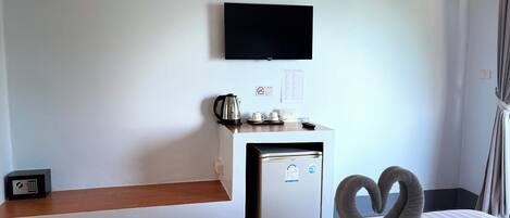 A hotel room with a bed, a small desk with a kettle, a television, and a refrigerator.