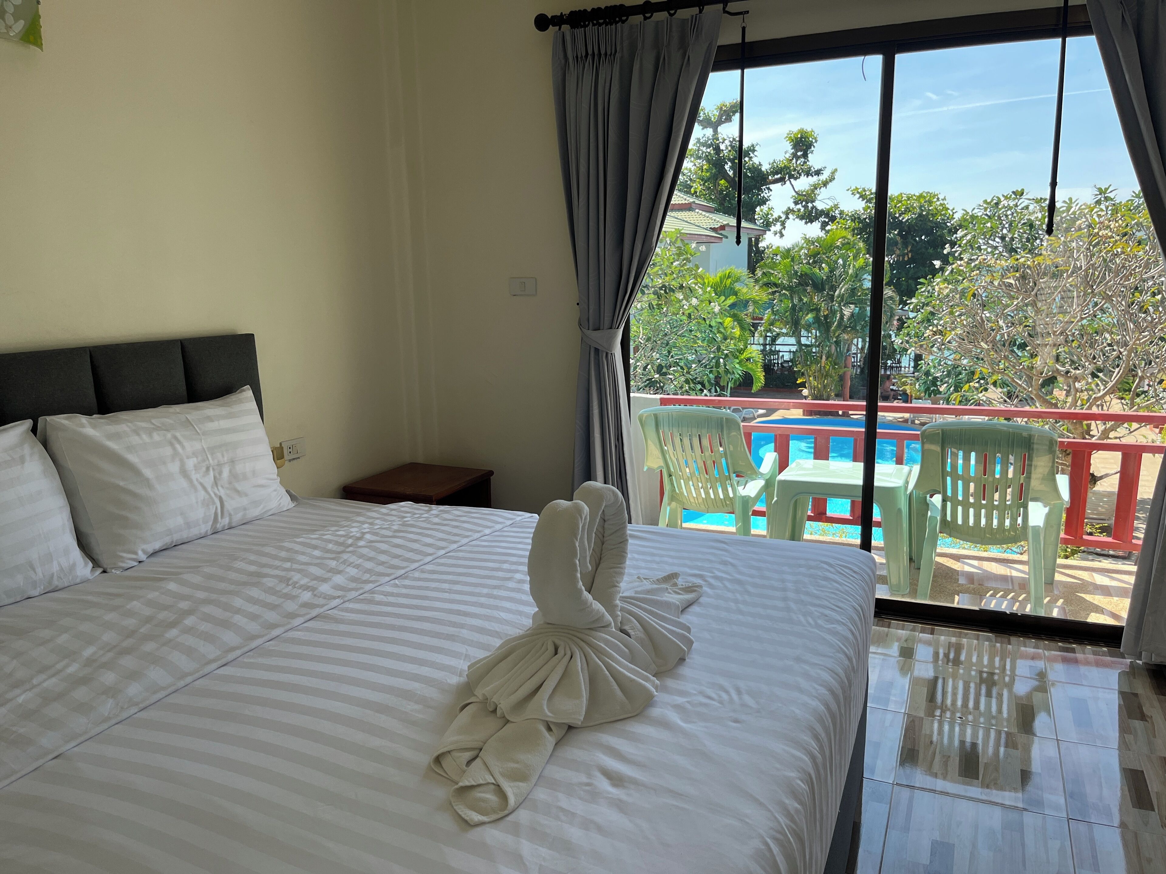 Une chambre d’hôtel avec un lit, une petite table et une vue sur une piscine et la verdure environnante.