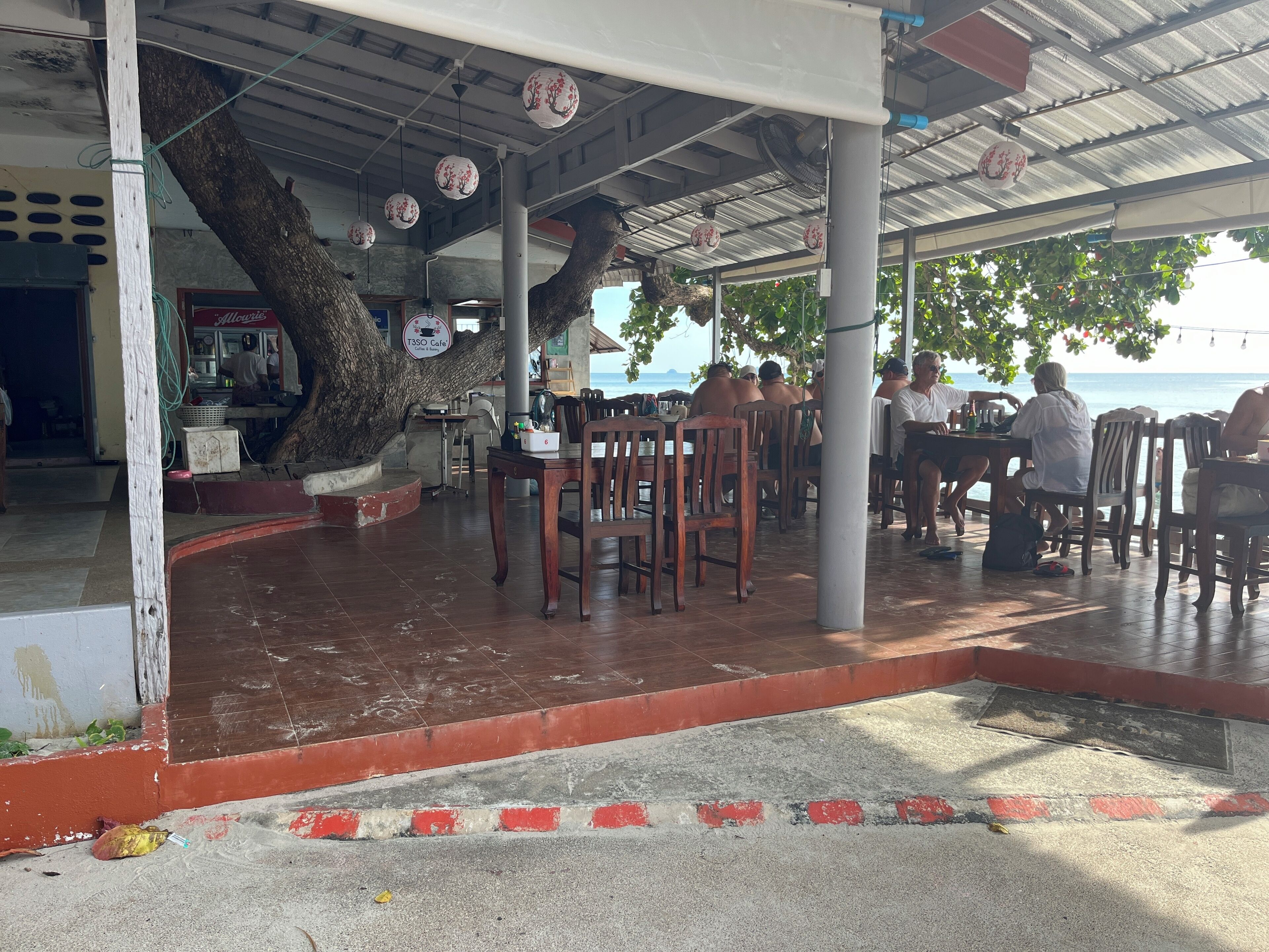 Un café al aire libre con mesas y sillas de madera, un árbol que da sombra y vista al mar.