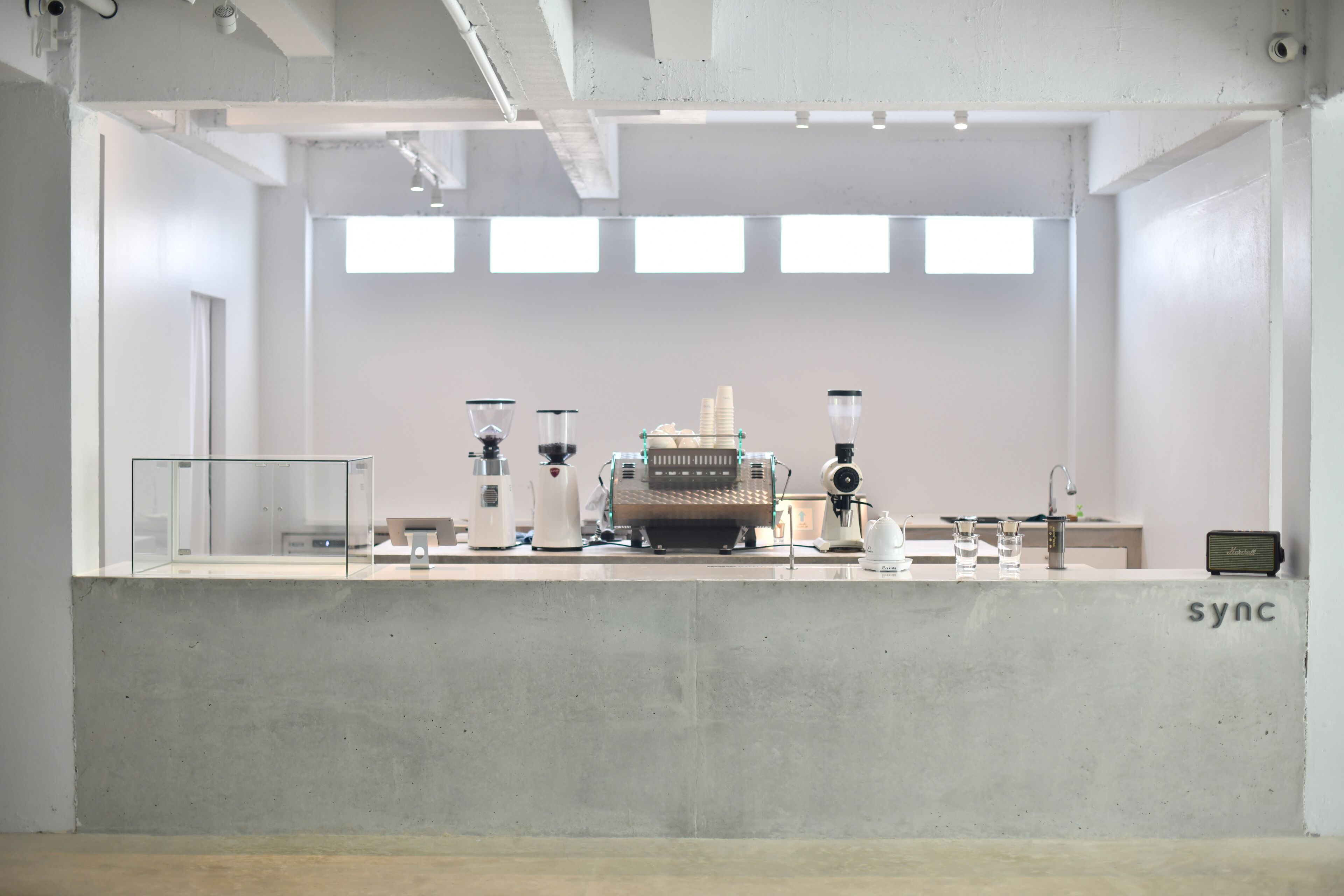 A minimalist interior with a concrete counter, various coffee-making equipment, and a small table with a radio.