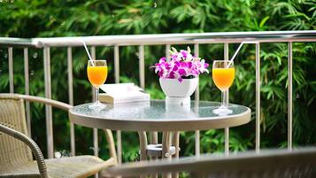 Une table ronde en verre, sur laquelle sont posées deux boissons et un vase de fleurs, est placée devant une rambarde en métal, avec une végétation luxuriante en arrière-plan.