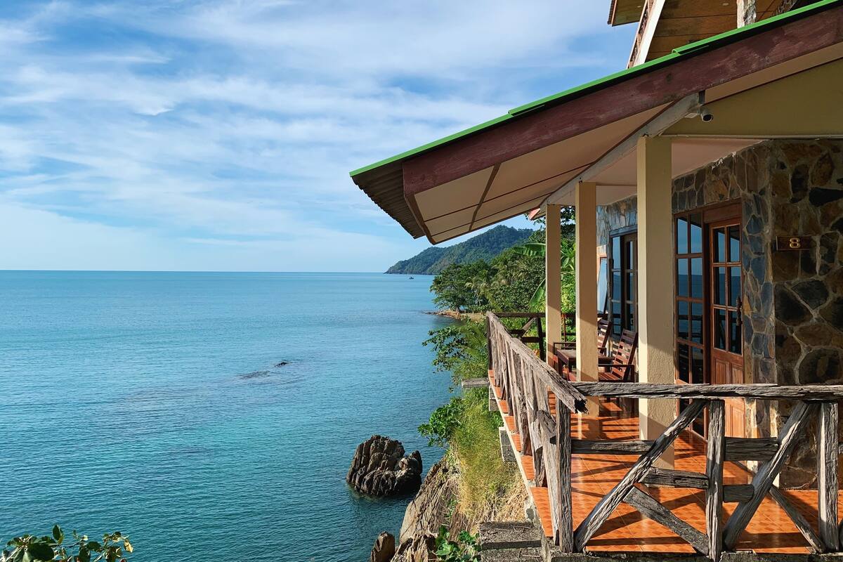 Ein Küstengebäude mit Steinaußenwand, einer Holzterrasse und Blick auf das Meer.