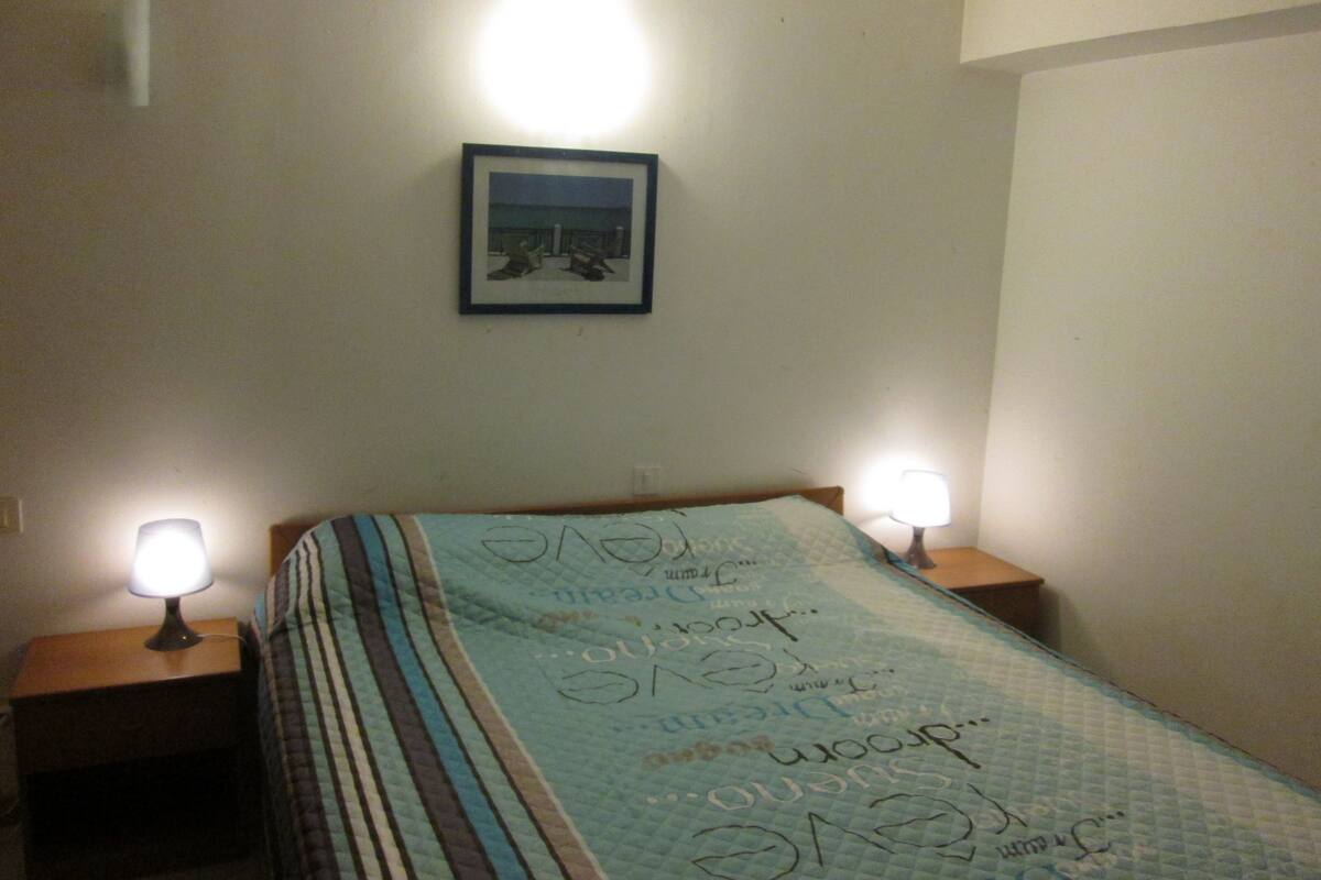 A bedroom with a bed, two bedside lamps, a framed picture on the wall, and a quilt with text.