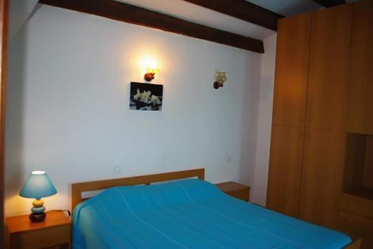A bedroom with a bed, a nightstand with a lamp, a wall-mounted light, and a small picture.
