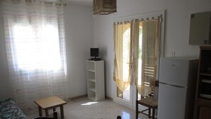 A small, well-lit room with a dining table, chairs, a television on a stand, a refrigerator, and a window with curtains. - Résidence Anna (Centuri)