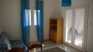 A room with a bed, a chair, a small table, a wooden cabinet, and a sliding glass door leading to an outdoor area. - Résidence Anna (Centuri)