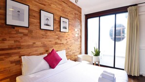 A modern hotel room with a wooden wall, a large bed with white bedding and a red pillow, a small round table with a plant, and a large window with curtains. - The Rommanee Boutique Guesthouse (Phuket)