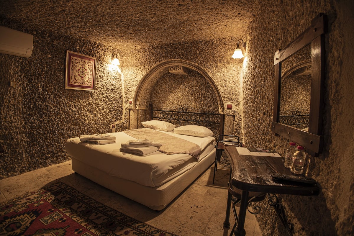 Un dormitorio con pared de piedra, una cama con cabecera, mesitas de noche, un espejo y una alfombra.