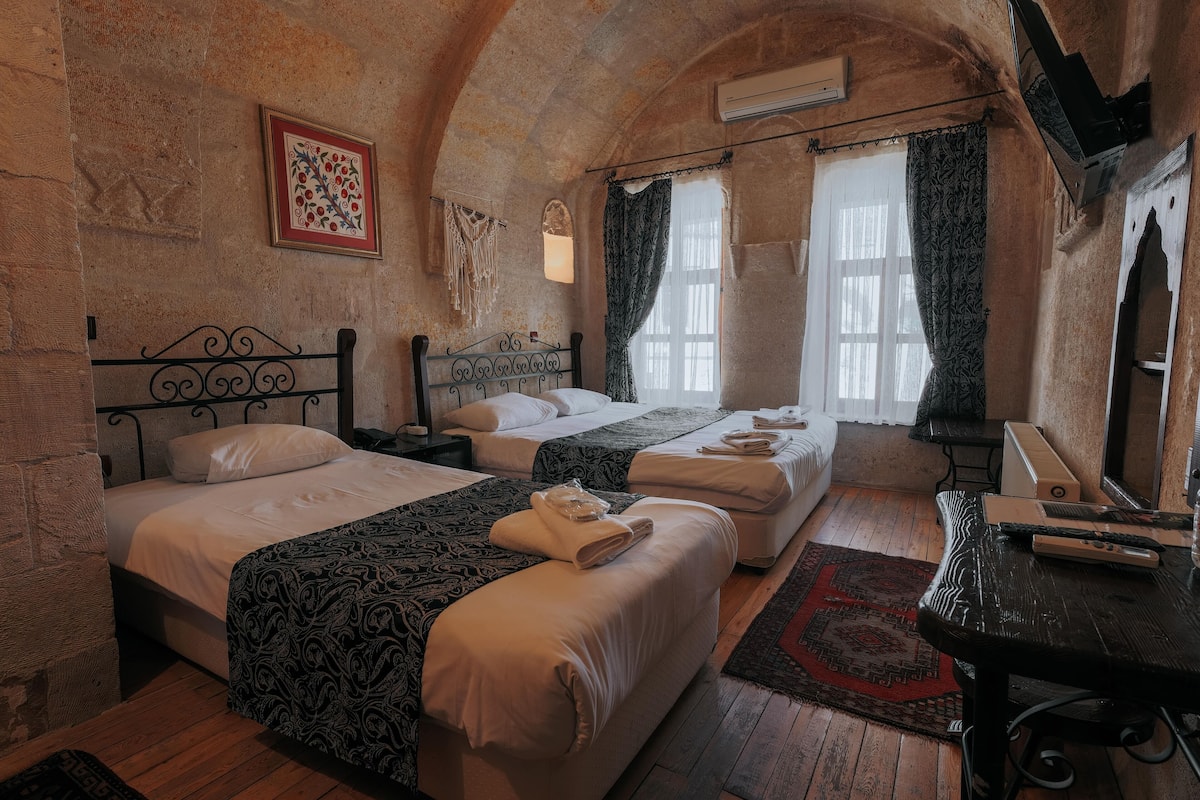 Habitación con dos camas, un escritorio y un espejo. Se observa un cuadro en la pared y una ventana con cortinas.