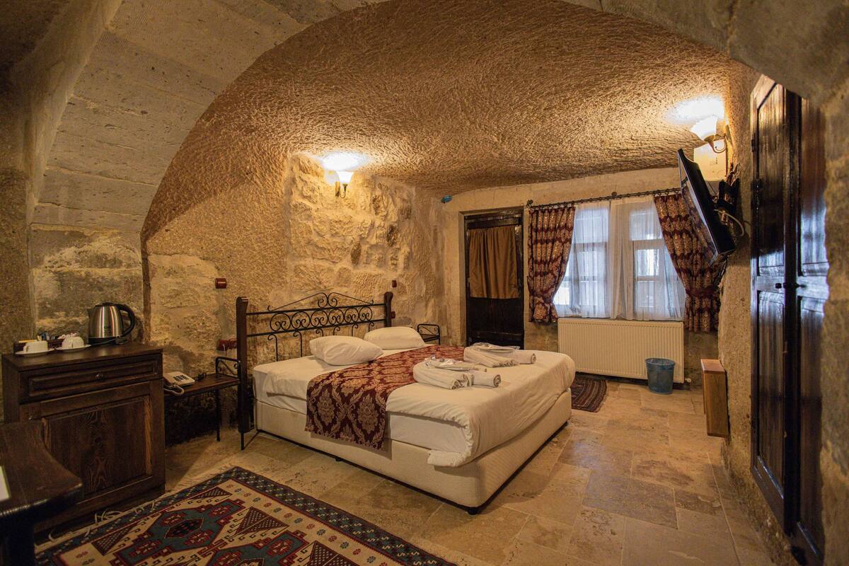 Un dormitorio con techo de piedra, una cama con ropa blanca, un aparador de madera, un radiador y una ventana con cortinas.