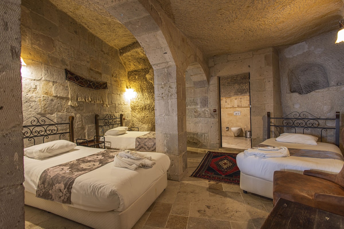 Una habitación de paredes de piedra con dos camas, una mesa pequeña y un banco.