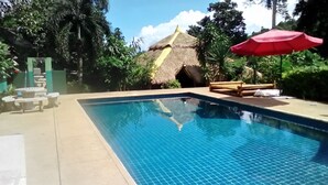 Una piscina con una cabaña de techo de paja, rodeada de vegetación y un paraguas rojo.