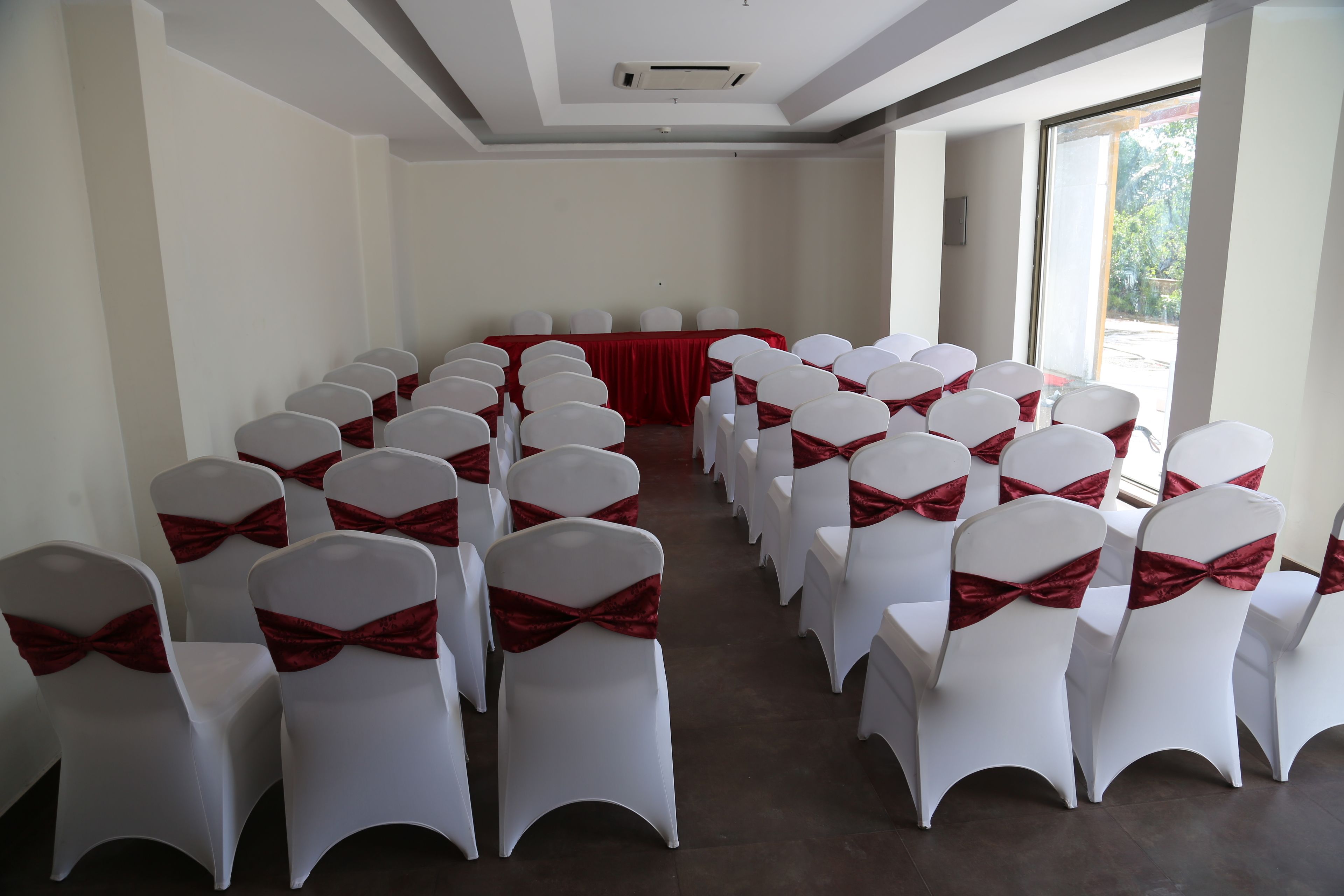 A conference room with rows of white chairs with red sashes, a red tablecloth-covered table, and large windows allowing natural light.