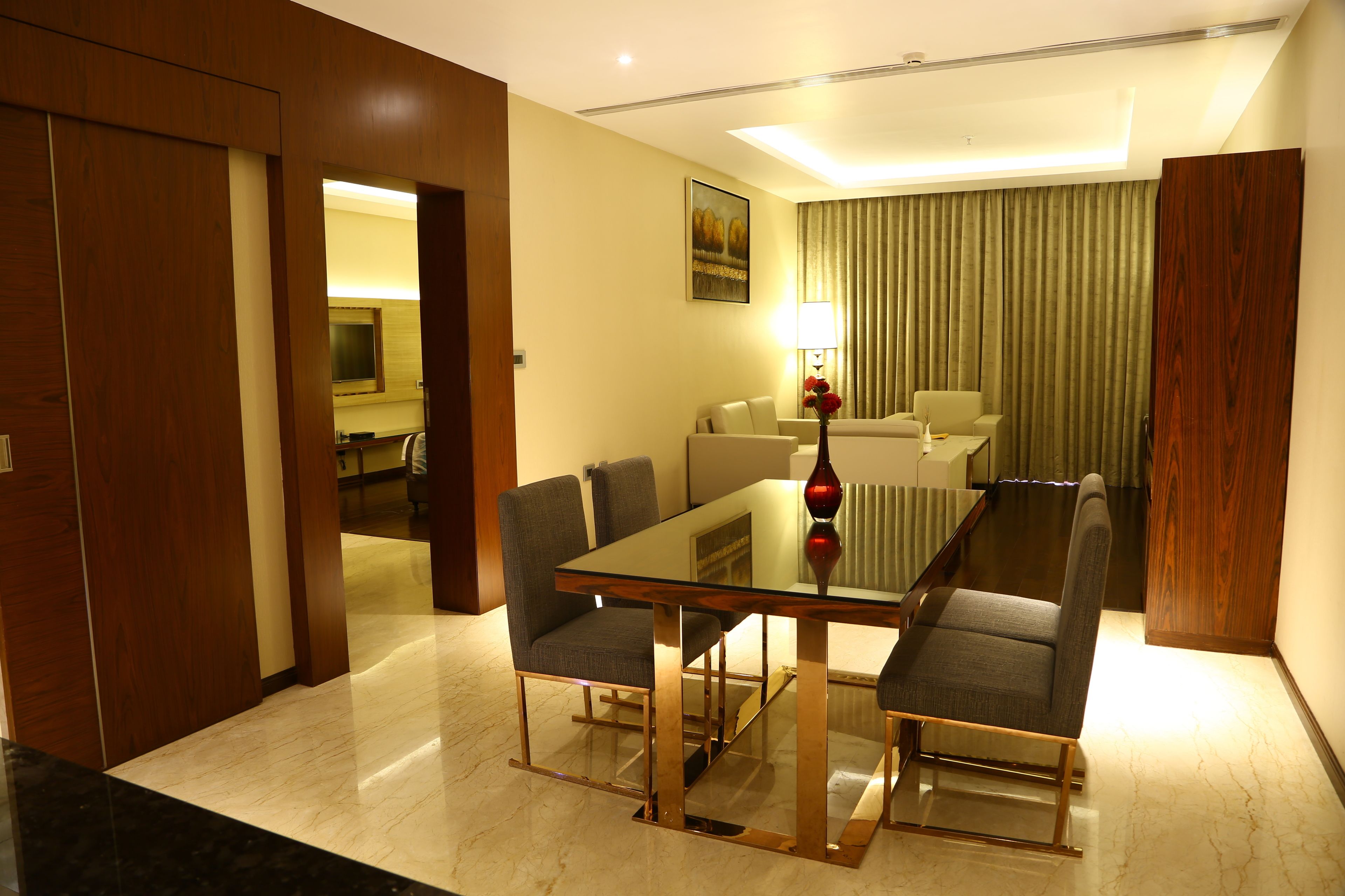 Habitación de hotel con zona de comedor que incluye una mesa de vidrio y sillas de estructura metálica, un sofá y una lámpara.