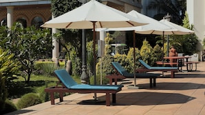 A patio area with lounge chairs and umbrellas, surrounded by greenery and a building with arched windows. - UVA Meridian bay resort and spa (Kundapur)