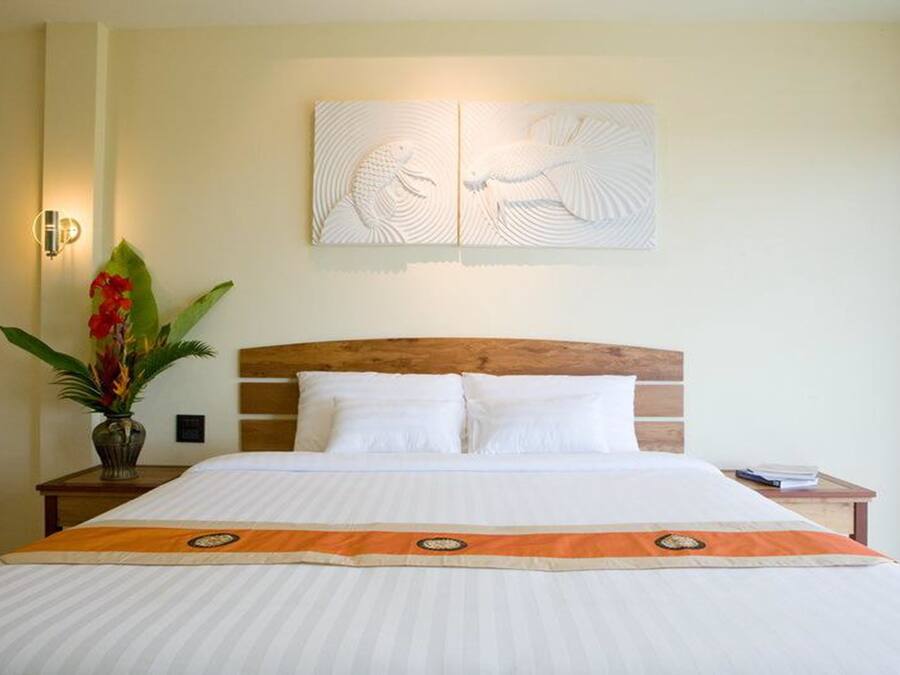 Una habitación de hotel con una cama grande, cabecera de madera, ropa de cama blanca, un adorno en la pared, una mesita de noche con un florero y una pequeña mesa con un libro.