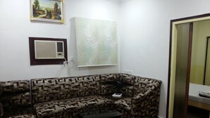 Una sala de estar con un sofá estampado, una mesita, un equipo de aire acondicionado y una ventana con una planta.
