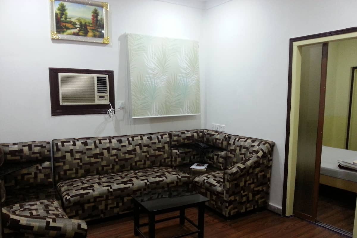 Una sala de estar con un sofá estampado, una mesita, un equipo de aire acondicionado y una ventana con una planta.