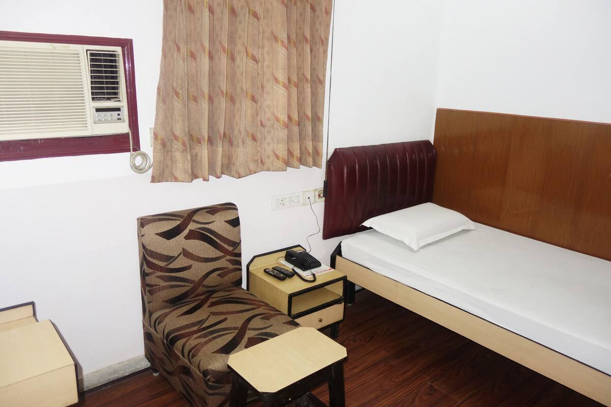 Una habitación pequeña y funcional con una cama individual, una silla, una mesita, una mesita de noche con teléfono y mando a distancia, y una ventana con equipo de aire acondicionado.
