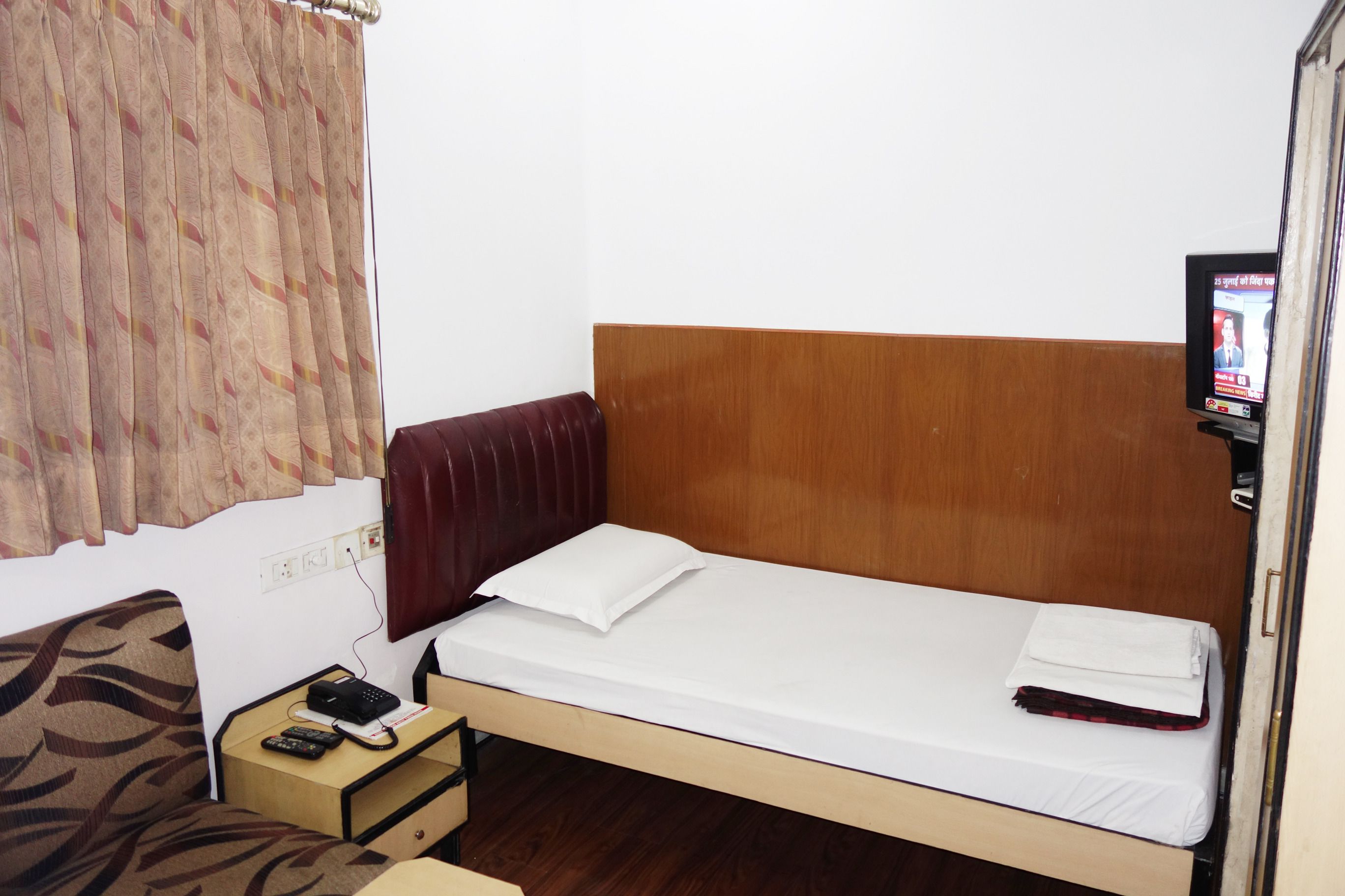 A single bed with a wooden headboard, a small bedside table with a remote control and a book, and a television on the wall.