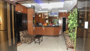 Zona de recepción de un hotel con un mostrador de madera, dos recepcionistas, un sofá, una mesita y una planta en maceta.