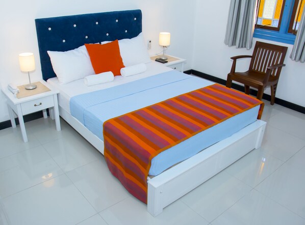 Una cama bien hecha con cabecera azul, almohadas blancas y manta a rayas. Hay dos mesitas de noche con lámparas y una silla de madera.