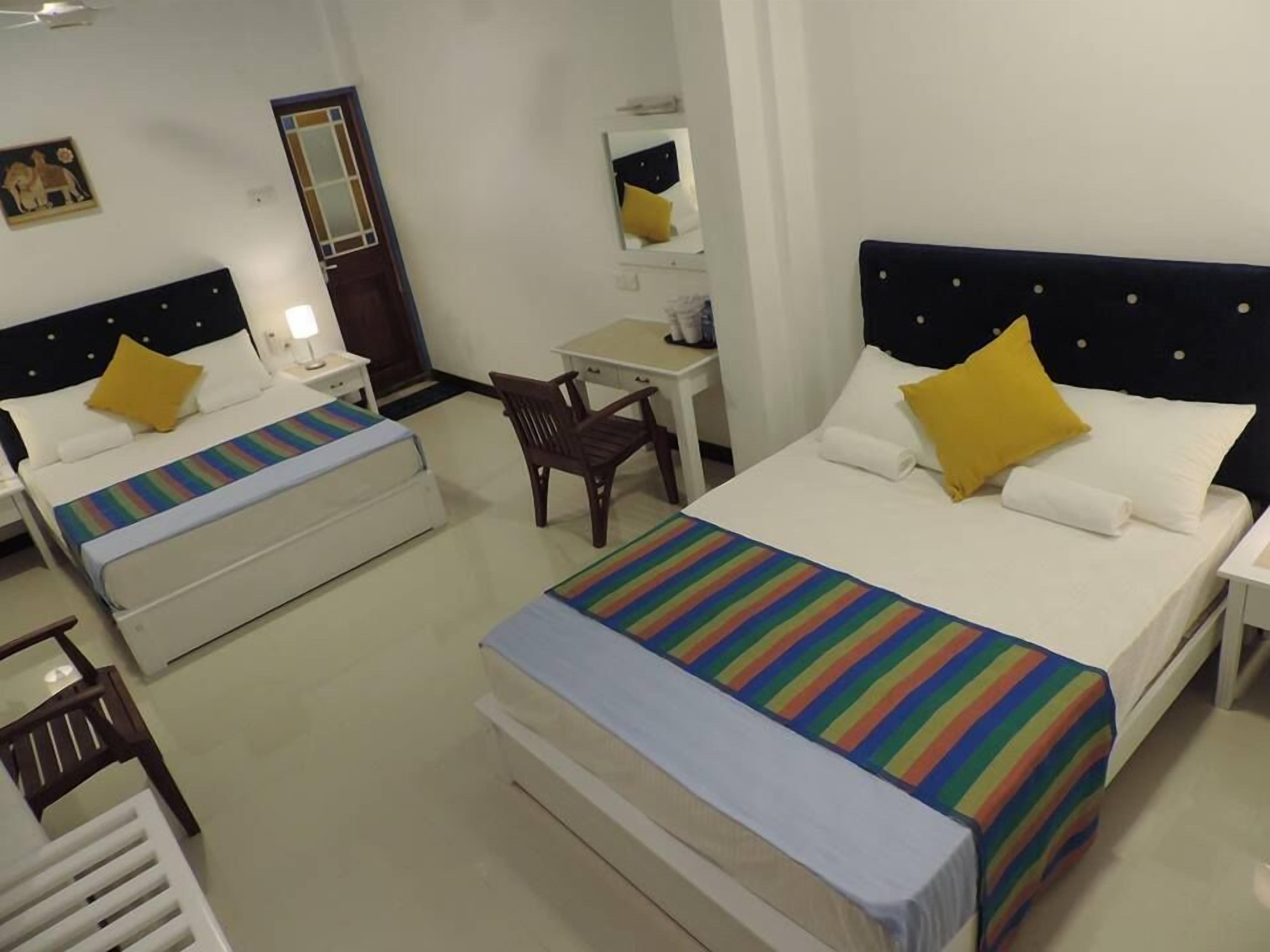 A hotel room with two single beds, each with a striped bedspread and a yellow pillow. There is a small desk with a chair, a mirror, and a nightstand with a lamp.
