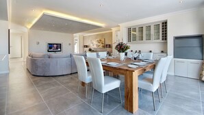 A modern dining area with a wooden table set for six, surrounded by white chairs. A grey sofa is positioned in the background, and a flat-screen TV is mounted on the wall. - Spatalla Holiday Homes (Kleinmond)