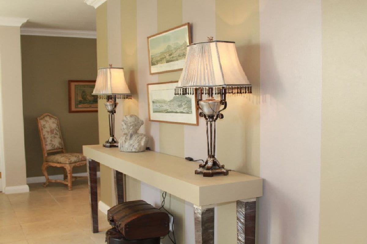 A hallway with a long, narrow console table flanked by two ornate lamps, a chair, and framed artwork on the wall.