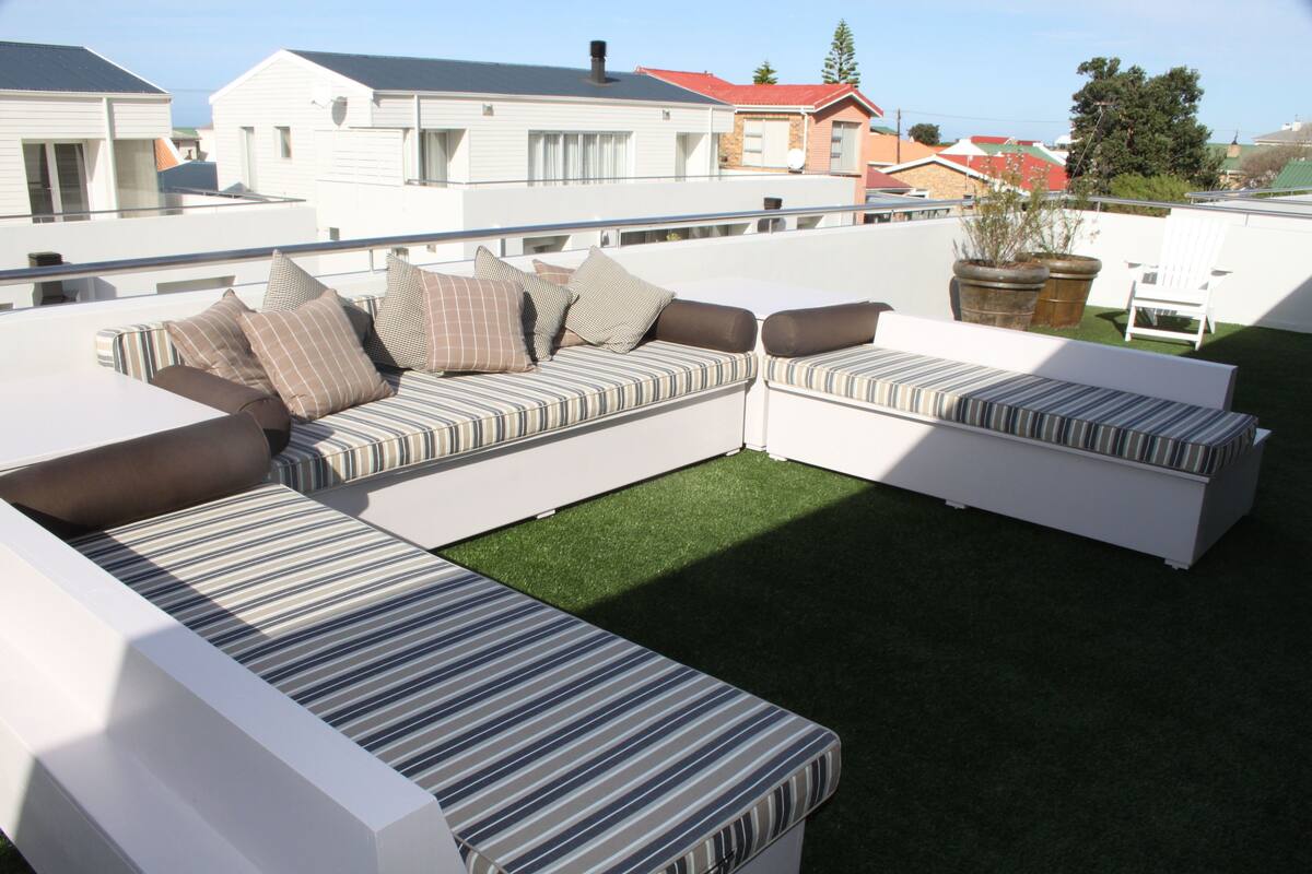 Terraza en la azotea con un sofá modular, una mesita pequeña, césped verde y barandilla blanca.