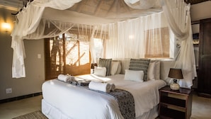 A bedroom with a canopy bed, a bedside table with a lamp, and a window with blinds. - Raptors Lodge (Hoedspruit)