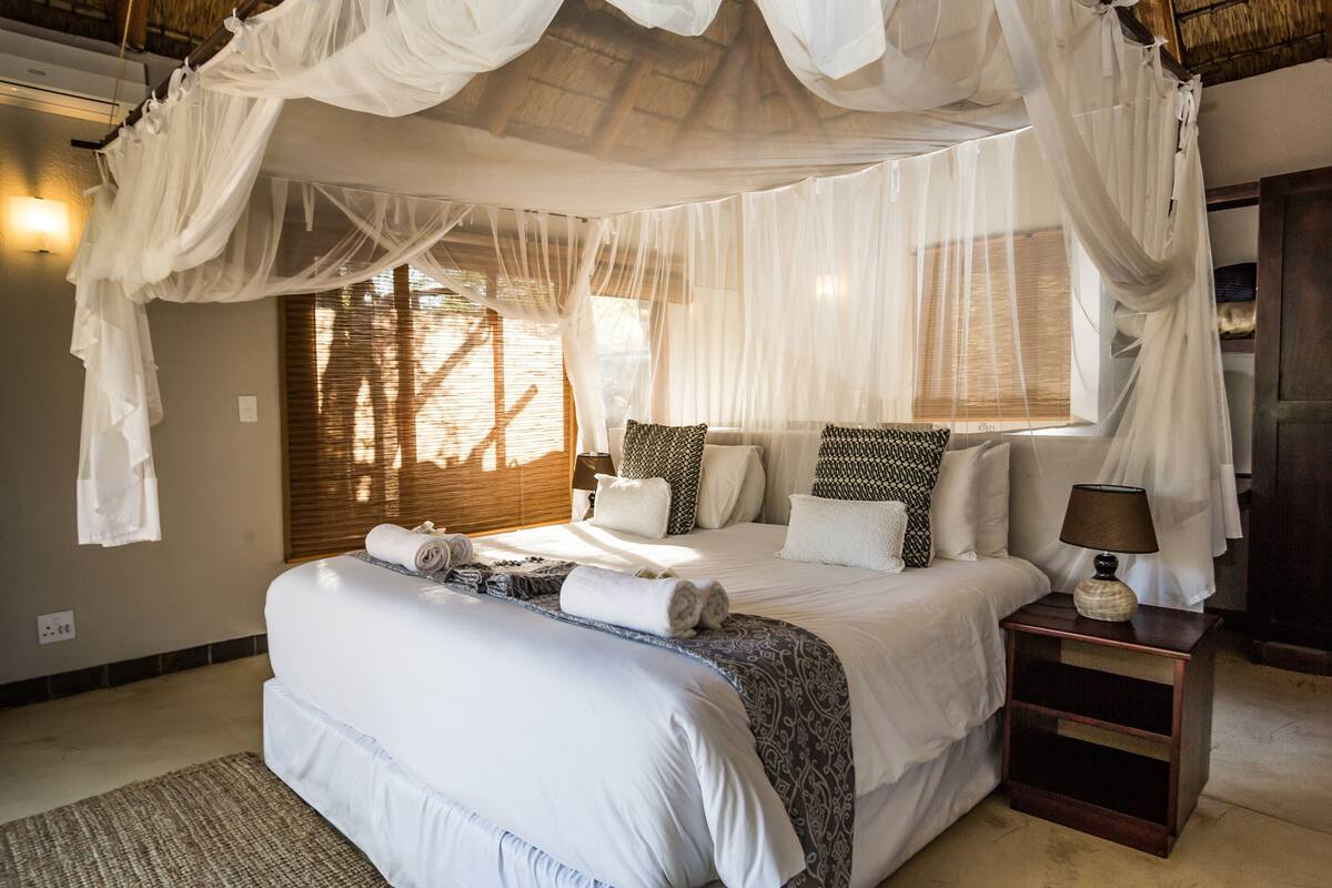 Un dormitorio con cama a dosel, una mesita de noche con lámpara y una ventana con persianas.