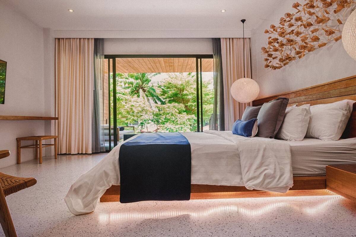 A modern bedroom with a large bed, wooden headboard, and a view of greenery outside.