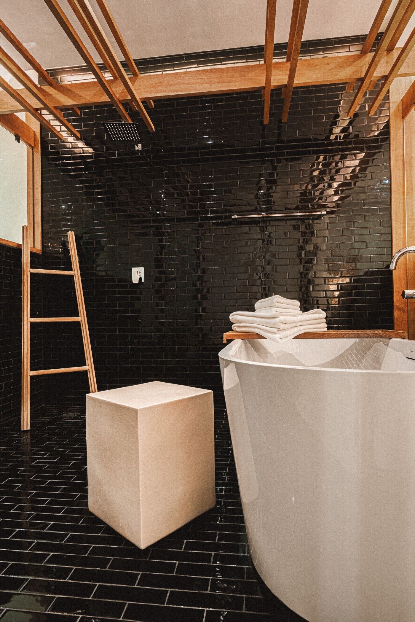 A modern bathroom with a black tiled wall, a wooden ladder, a large bathtub, and a stack of white towels on a wooden shelf.