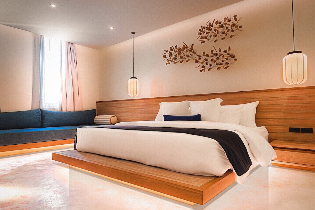 A modern bedroom with a wooden headboard, a large bed with white and dark pillows, a bench with cushions, and a wall art piece.