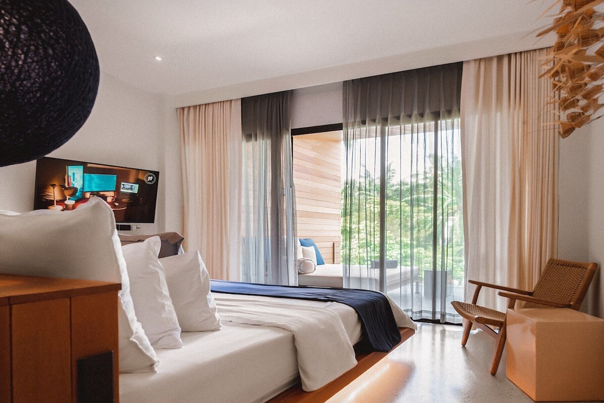 A modern hotel room with a bed, a TV, a seating area, and a view of greenery outside.