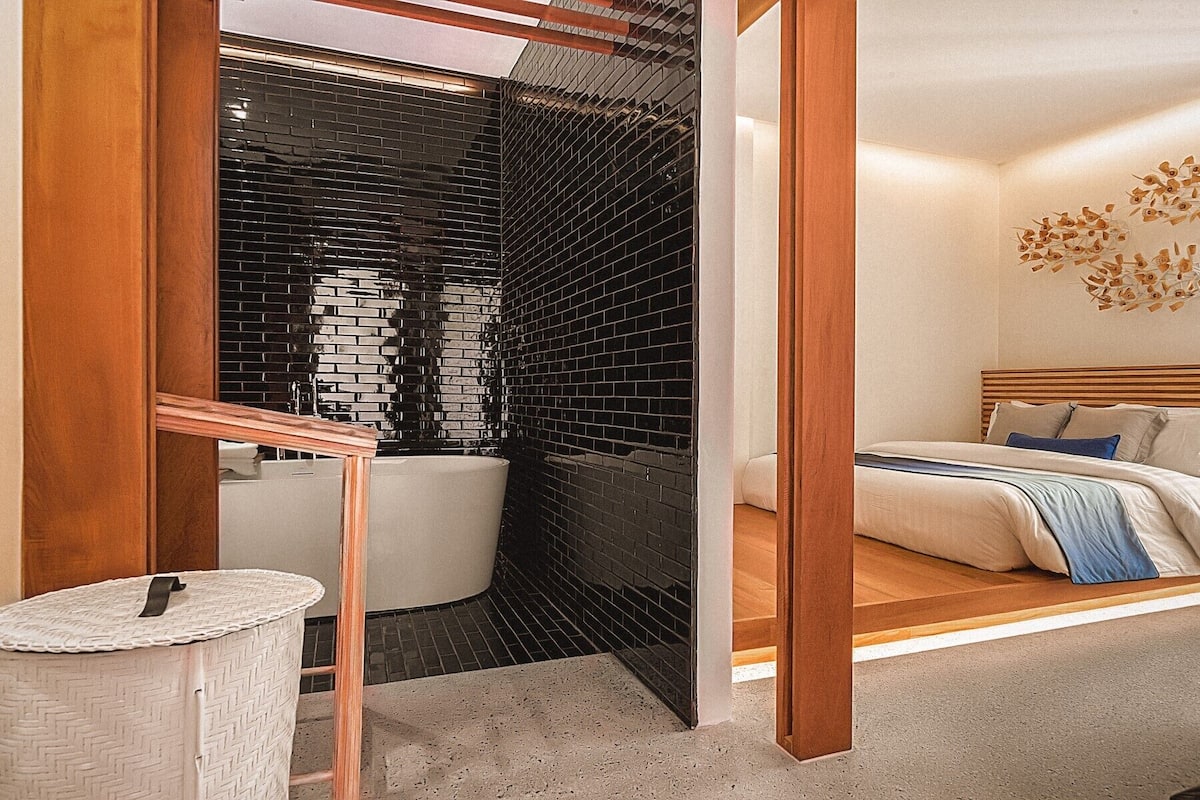 A modern bathroom with a black tiled wall, a bathtub, and a wooden-framed door leading to a bedroom with a bed and a wall-mounted light fixture.