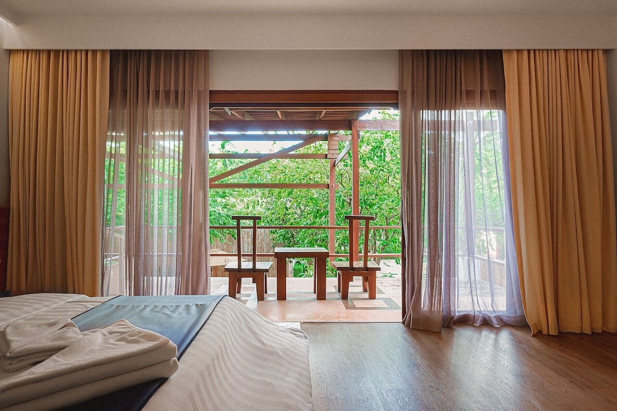 A hotel room with a bed, a wooden table, and a view of a garden through large windows.