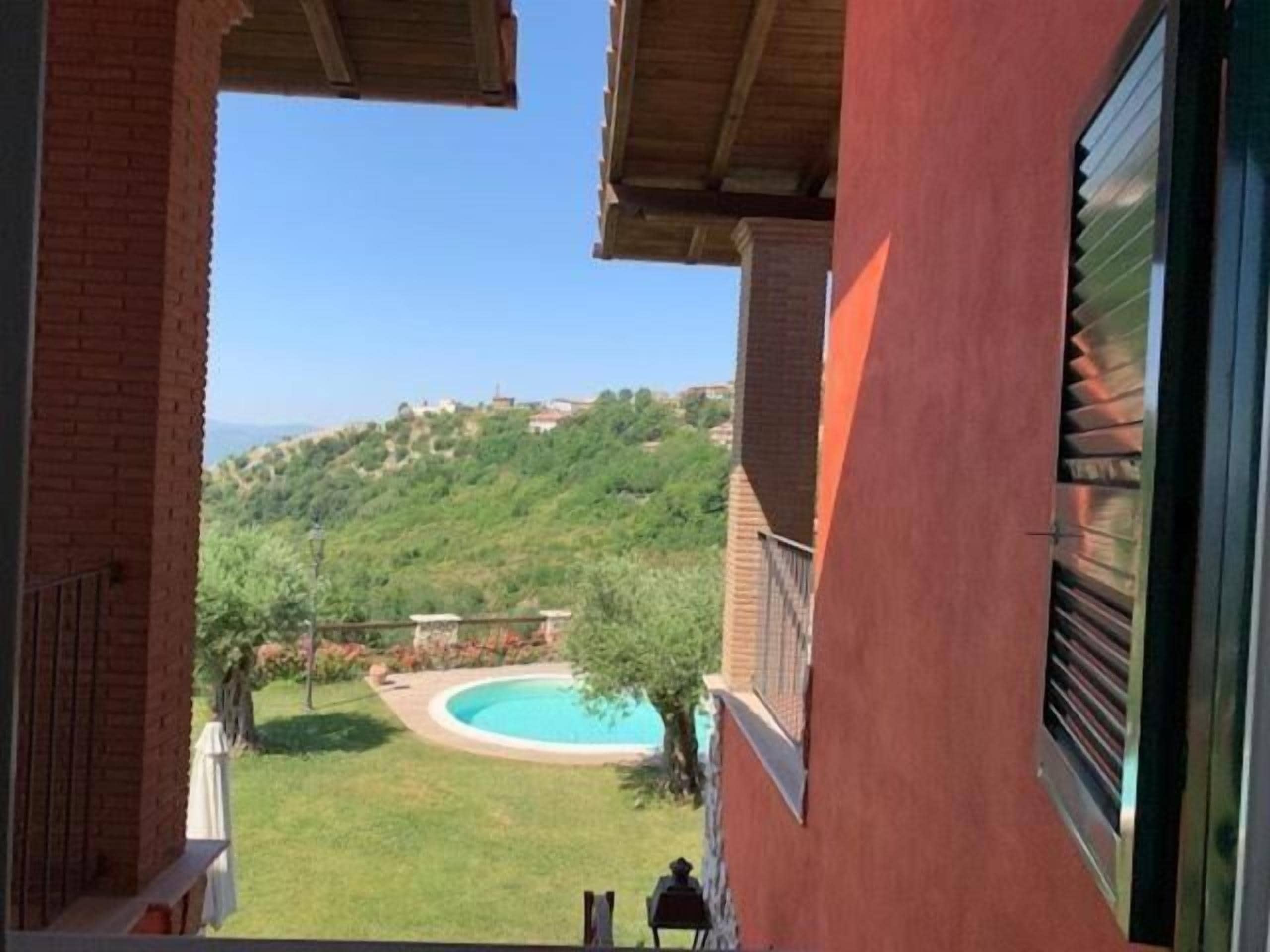 A view from a balcony overlooking a swimming pool, a grassy area, and a hilly landscape.