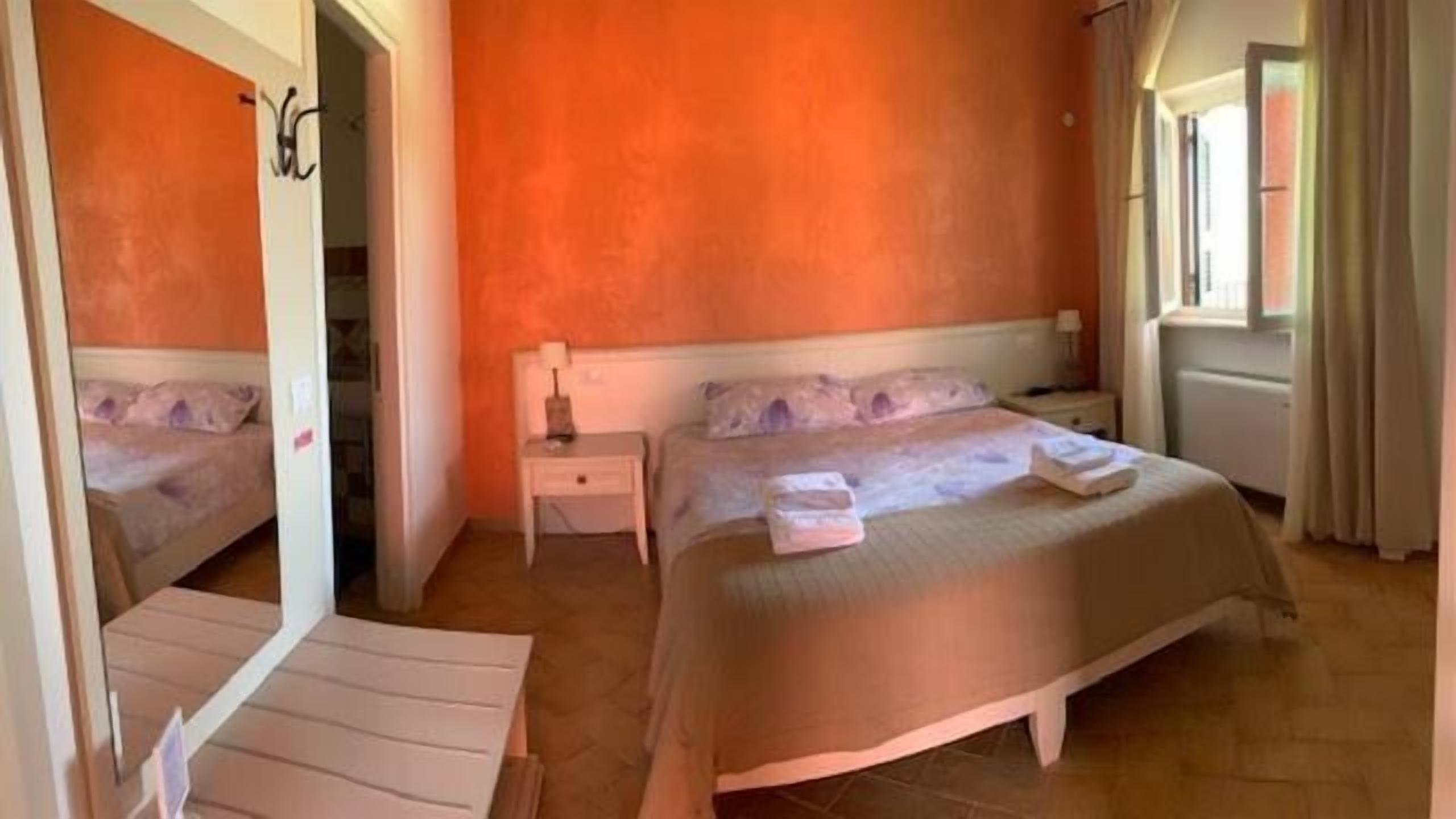 A hotel room with a large bed, a bedside table, and a nightstand. There is a window with curtains and a door leading to another room.