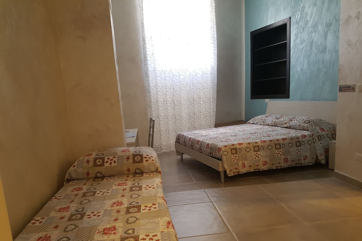 A small, well-lit room with two beds, a window with patterned curtains, and a wall-mounted TV.