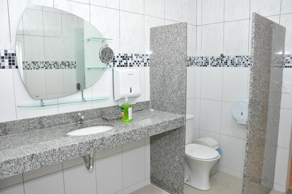A bathroom with a granite countertop sink, a glass soap dispenser, a toilet, and a shower area. - Hotel Nacional (Rondonopolis)