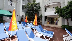 Zona de piscina con sillas reclinables azules y sombrillas rojas y amarillas, rodeada por un edificio blanco con balcones.